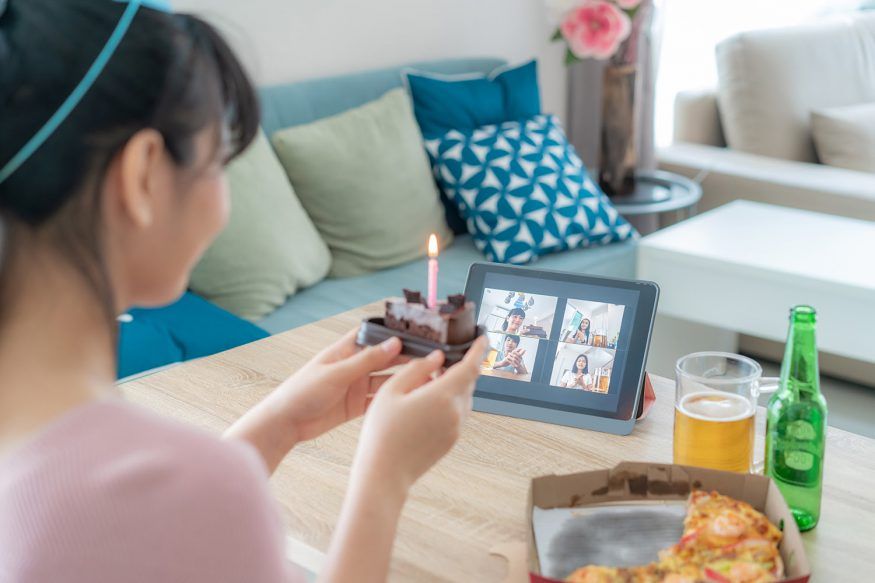 A woman holds a piece of cake in front of a tablet, celebrating a virtual birthday party